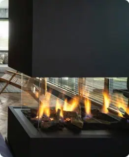 A close-up shot of a modern, multi-sided fireplace with a black frame and a glass enclosure. The realistic-looking flames are burning over ceramic logs. In the blurred background, a room with a wooden chair and blinds is visible.