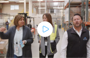 A video thumbnail showing three warehouse workers walking and talking in a warehouse aisle. A woman on the left, with brown hair and wearing a gray cardigan, gestures with her hand as she speaks. A woman in the center wears a yellow safety vest, and a man on the right, with short hair and a black vest, listens intently. A forklift is visible in the background. A play button icon is overlaid on the image.