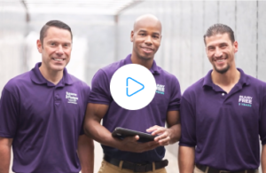 A video thumbnail image featuring three smiling men wearing purple polo shirts. The man in the center is holding a tablet and a play button icon is overlaid on the image, suggesting it is a video still. The other two men are flanking him, all looking at the camera.