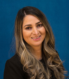 Professional headshot of a smiling woman with long wavy hair, wearing a black blazer, against a solid blue background.