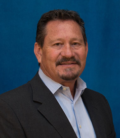 Professional headshot of a middle-aged man with short dark hair and facial hair, wearing a dark suit jacket over a light blue dress shirt, smiling slightly against a blue backdrop.