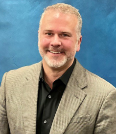 Smiling man in a grey checkered blazer and black shirt posing in front of a blue background.